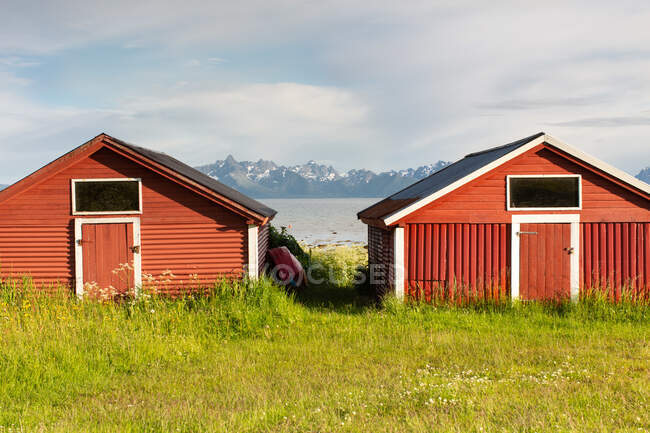 Red wooden buildings located on green meadow on shore with rocky mountains and cloudy sky in background in sunny day in countryside — Stock Photo