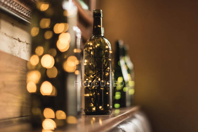 Складені прозорі скляні пляшки з блискучими світлими гірляндами всередині, поміщені в ряд на дерев'яній полиці біля стіни — стокове фото