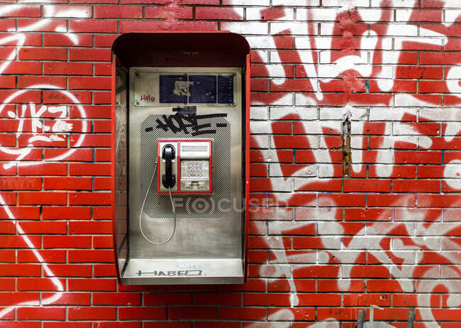 Городской телефон-автомат висит на стене из красного кирпича с белыми граффити на городской улице — стоковое фото