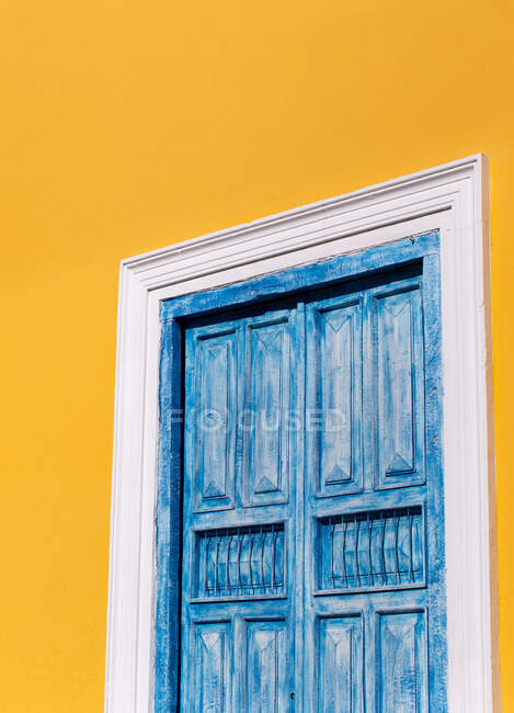 Shut window with shabby blue wooden shutters on bright yellow wall of building in sunshine — Stock Photo
