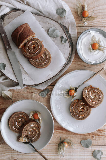 Сверху нарезанный вкусный домашний торт из сладкого ролла со взбитыми сливками и сушеные цветы Физиалиса, подаваемые на тарелке на деревянном столе с ингредиентами — стоковое фото