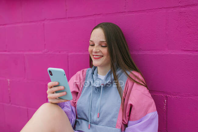 Adolescente alla moda seduto vicino al muro rosa con smartphone — Foto stock