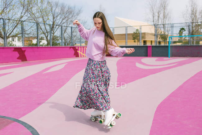 Encantadora joven hembra en falda colorida y sudadera lila de pie sobre patines de cuatro ruedas en el patio de recreo rosa en el día de primavera en la ciudad - foto de stock