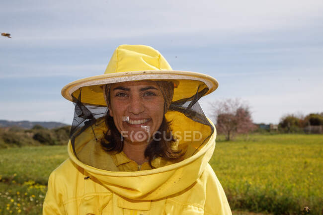 Positive female beekeeper in yellow protective costume and mask smiling and looking at camera while standing in green field in apiary in sunny summer day — Stock Photo