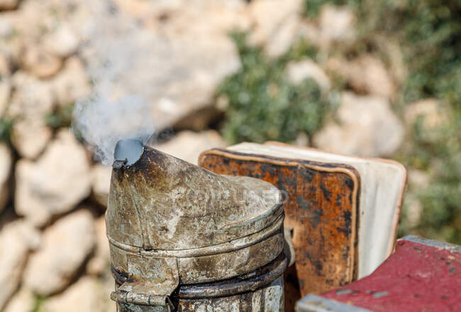 Primer plano del fumador de abejas metálicas oxidadas erosionadas con humo colocado sobre un fondo natural borroso en el colmenar - foto de stock