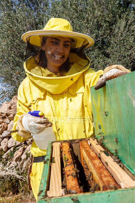 Apicultor fêmea feliz em desgaste protetor amarelo usando dispensador enquanto pulveriza colmeia com abelhas em apiário — Fotografia de Stock