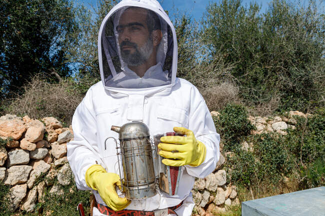 Серьезный профессиональный пчеловод в защитном костюме и перчатках держит курильщика пчел и смотрит в сторону, стоя на пасеке в летний день — стоковое фото