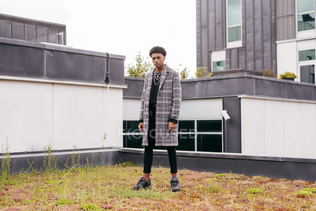 Серьезный молодой афро-американский модный парень в модном клетчатом пальто и узких штанах, смотрящий в камеру, стоя напротив современных зданий в городе — стоковое фото