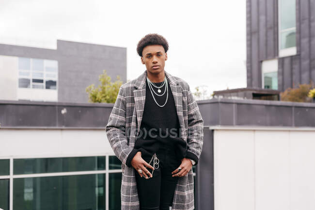 Sérieux jeune homme afro-américain à la mode en manteau à carreaux à la mode et pantalon serré regardant la caméra tout en se tenant contre les bâtiments modernes de la ville — Photo de stock