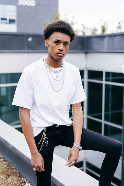 Молодий афроамериканець у модний одяг, сталеві ланцюжки та браслет, який дивиться на камеру, стоячи біля сучасного будинку в місті. — стокове фото