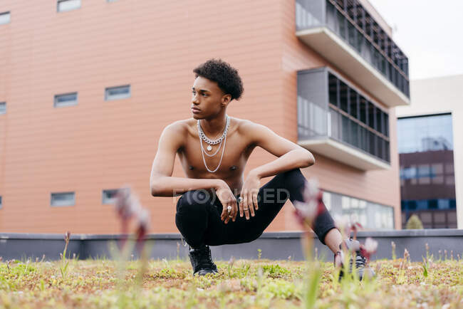 Уверенный молодой афроамериканец с цепными ожерельями на голом туловище, одетый в узкие брюки и модные сапоги, приседающие на улице с современным зданием на заднем плане — стоковое фото