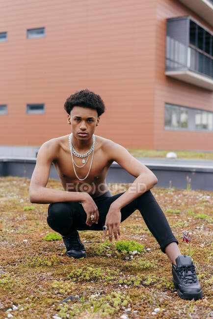 Confident young African American male with chain necklaces on naked torso dressed in tight pants and trendy boots squatting on street with modern building in background — Stock Photo