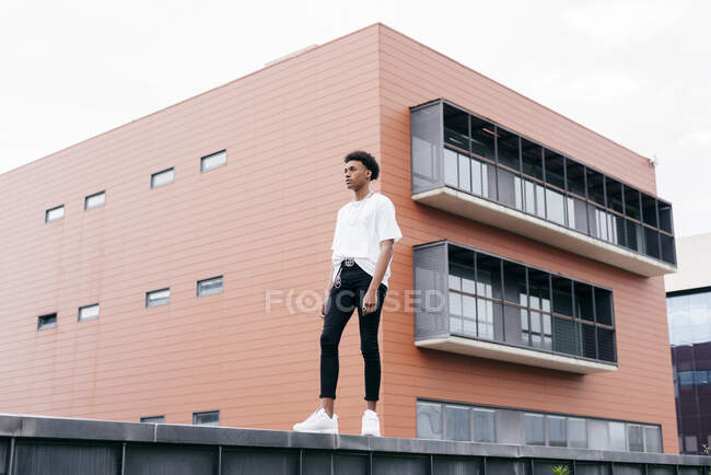 From below side view of young stylish African American male looking away in white shirt and tight pants with white sneakers standing on fence against pink city building — Stock Photo