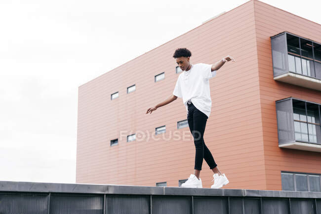 Dal basso vista laterale del giovane maschio afroamericano alla moda guardando lontano in camicia bianca e pantaloni stretti con scarpe da ginnastica bianche in piedi sulla recinzione contro l'edificio della città rosa — Foto stock