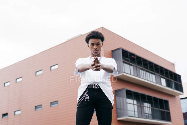Low angle of stylish independent young African American man in trendy outfit with steel accessories looking at camera gesturing a gun with hands while standing against pink building on city street — Stock Photo