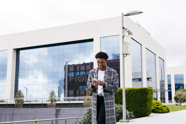 From below of happy young African American male student in stylish outfit browsing smartphone while standing on city street near modern building — Stock Photo