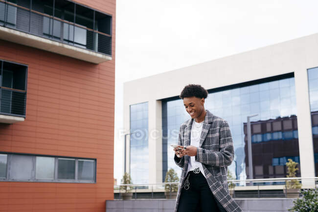 From below of happy young African American male student in stylish outfit browsing smartphone while standing on city street near modern building — Stock Photo