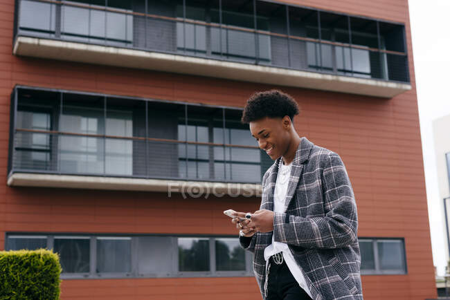 Von unten Seitenansicht eines glücklichen jungen afroamerikanischen männlichen Studenten in stylischem Outfit, der in der Nähe eines modernen Gebäudes auf der Straße steht und sein Smartphone durchsucht — Stockfoto