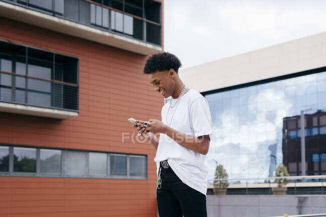 From below side view of happy young African American male student browsing smartphone while walking on city street near modern building — Stock Photo