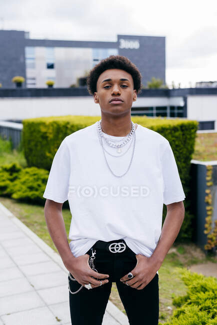 Young black curly haired male teenager in stylish clothes with chains looking at camera while standing on city street with green plants and buildings in background — Stock Photo