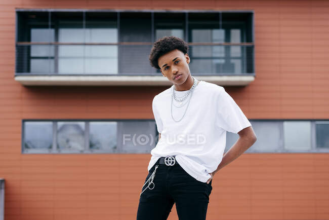 Молодий чорношкірий кучерявий чоловічий підліток у стильному одязі з ланцюгами, дивлячись на камеру, стоячи на міській вулиці з будівлями на задньому плані — стокове фото