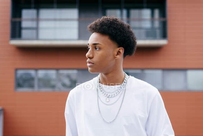 Jovem preto encaracolado adolescente masculino em roupas elegantes com correntes olhando para longe, enquanto em pé na rua da cidade com edifícios no fundo — Fotografia de Stock