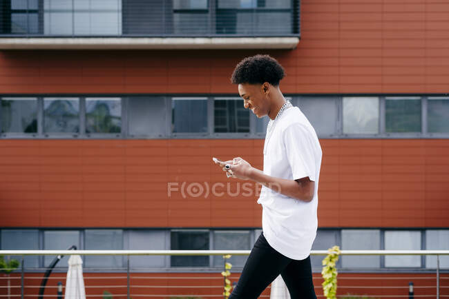 A partir de baixo vista lateral do jovem estudante afro-americano feliz que navega smartphone enquanto caminha na rua da cidade perto do edifício moderno — Fotografia de Stock