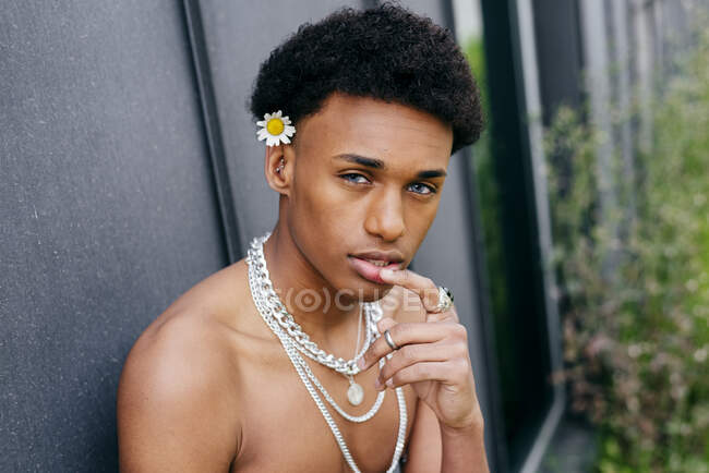 Seitenansicht verführerischer junger schwarzer Teenager mit nacktem Oberkörper und Halsketten, die Blume im Mund halten und in die Kamera schauen, während er vor grauer Wand steht — Stockfoto