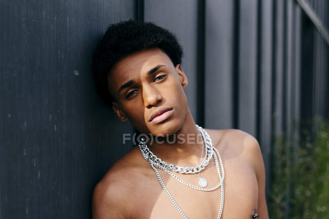 Verführerischer junger schwarzer Teenager mit nacktem Oberkörper und Halsketten, Blume im Mund und Blick in die Kamera, während er vor grauer Wand steht — Stockfoto