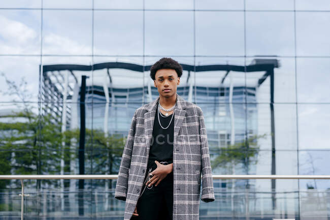 Selbstbewusstes junges afroamerikanisches Teenie-Model im trendigen Schottenmantel und stilvollen Accessoires, das vor einem modernen Gebäude mit Glaswand in die Kamera blickt — Stockfoto