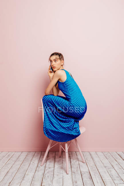 Vue latérale de la jeune femme accroupie sur le dessus d'une chaise regardant loin tout en parlant sur le téléphone mobile sur fond rose — Photo de stock