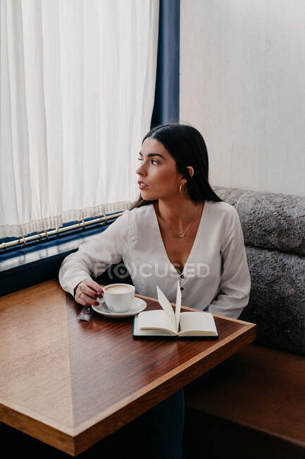Mujer morena con el pelo largo bebiendo café en un bar con un libro al lado - foto de stock
