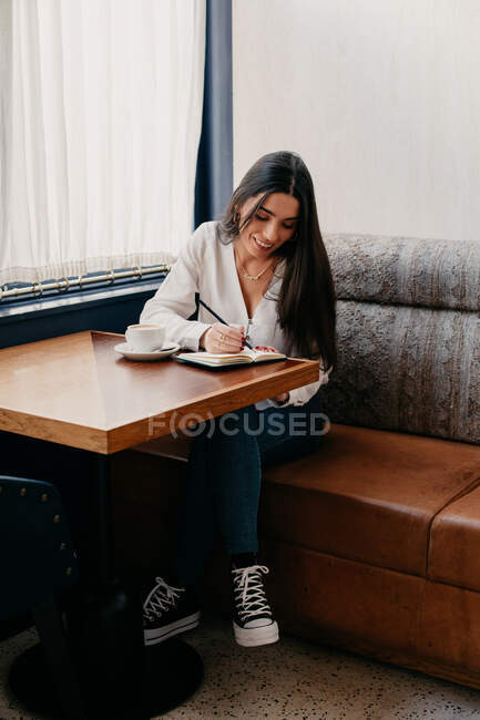 Mulher morena escrevendo em um caderno enquanto toma café em um bar — Fotografia de Stock