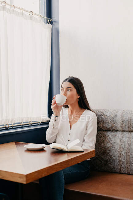 Brünette Frau mit langen Haaren trinkt Kaffee in einer Bar mit einem Buch daneben — Stockfoto