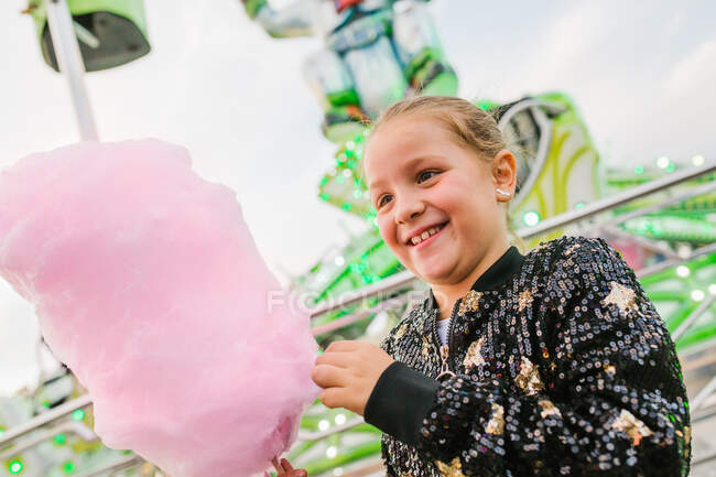 From below delighted girl smiling and eating sweet candyfloss while standing at funfair — Stock Photo