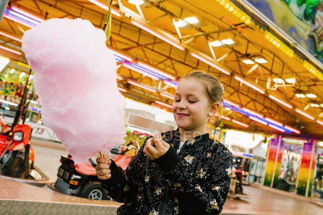 Chica encantada sonriendo y comiendo hilo de azúcar dulce, mientras que de pie en la feria - foto de stock
