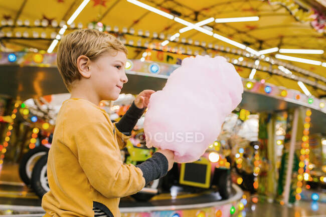 Vista lateral del niño encantado sonriendo y comiendo hilo de azúcar dulce mientras está de pie en la feria - foto de stock