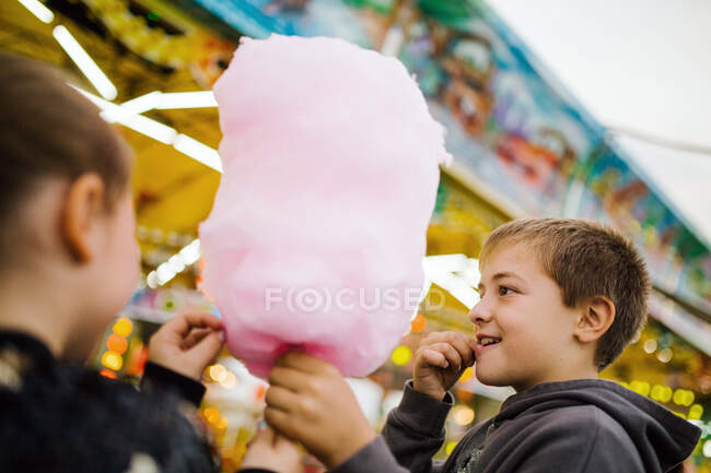 Vista lateral de la niña y los hermanos niños encantados sonriendo y comiendo hilo de azúcar dulce mientras está de pie en la feria - foto de stock