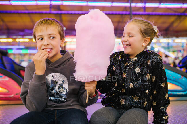 Deleitado niña y niños hermanos sonriendo y comiendo hilo de azúcar dulce mientras se sienta en la feria - foto de stock