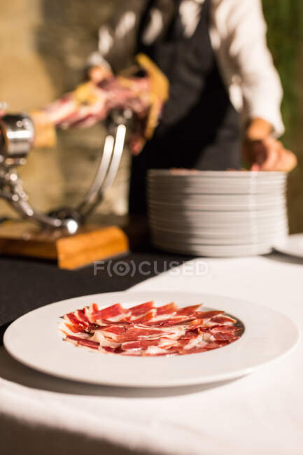 Crop unrecognizable person hand on apron cutting a whole dry-cured ham leg on a black background — Stock Photo