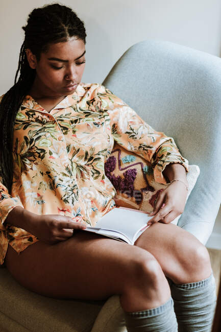 De cima de calma jovem étnica feminina em roupas casuais sentado na cadeira e ler livro enquanto passa o tempo livre em casa — Fotografia de Stock