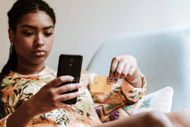 Jeune femme ethnique en tenue décontractée assise sur une chaise à la maison et effectuant une transaction en ligne avec smartphone et carte de crédit — Photo de stock