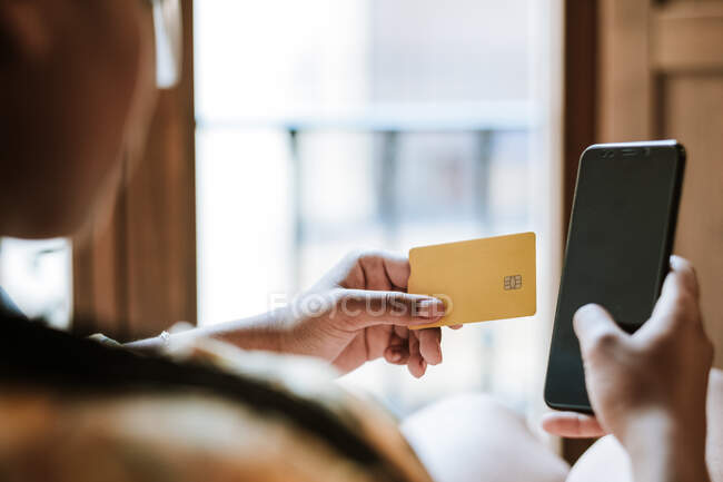 Vue arrière de la culture anonyme ethnique femelle avec carte de crédit et smartphone effectuer des achats en ligne tout en étant assis à la maison — Photo de stock