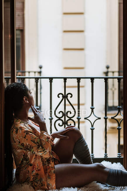 Vue latérale d'une femme ethnique silencieuse et rêveuse en tenue décontractée assise sur un balcon et regardant ailleurs tout en se reposant à la maison pendant le week-end — Photo de stock