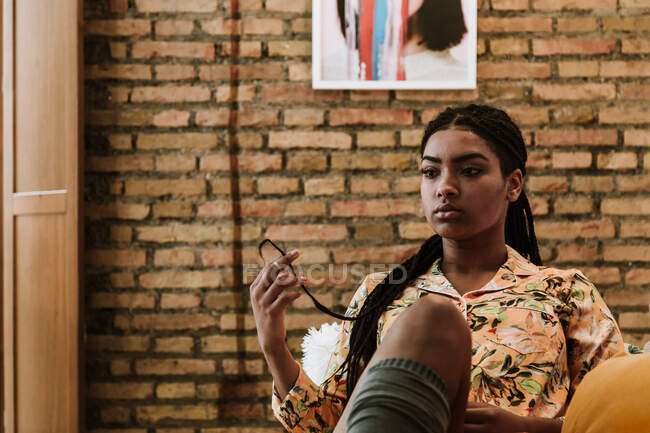 Молода афроамериканська жінка в повсякденному одязі сидить на дивані і думає про проблеми, проводячи час у сучасній квартирі з цегляною стіною. — стокове фото