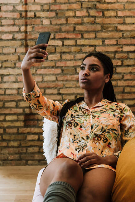 Joven mujer afroamericana seria en traje casual tomando selfie con teléfono inteligente mientras está sentado en la silla contra la pared de ladrillo en apartamento moderno - foto de stock