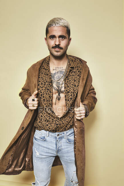 Modèle masculin à la mode avec des tatouages portant un manteau tendance sur une chemise léopard et un jean debout sur un fond beige et regardant la caméra — Photo de stock