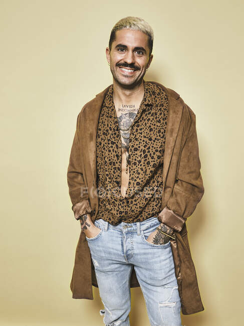 Весела модна чоловіча модель з татуюваннями у модному пальто над сорочкою леопарда та джинсами, що стоять на бежевому фоні та дивляться на камеру — стокове фото