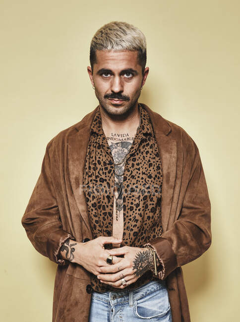 Modelo masculino de moda con tatuajes con abrigo de moda sobre camisa de leopardo y jeans de pie sobre fondo beige y mirando a la cámara - foto de stock