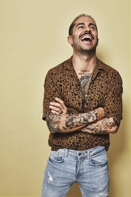 Весела модна чоловіча модель з татуюваннями у модній сорочці з леопардом та джинсах, що стоять на бежевому фоні та дивляться вбік — стокове фото